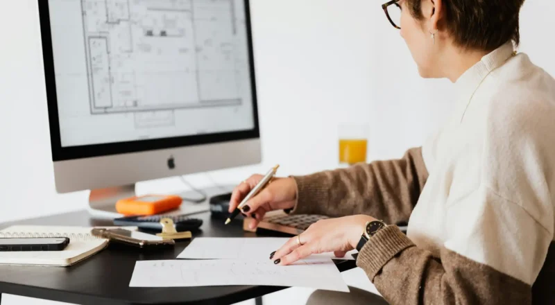 Pessoa trabalhando em projeto de arquitetura com computador, papel e materiais de escritório em mesa de trabalho.