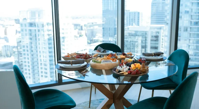 Mesa de jantar de vidro com pratos variados de comida ao lado de uma janela grande, com vista para prédios altos de uma cidade moderna.