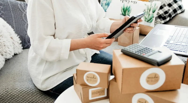Pessoa realizando gerenciamento de pedidos de produtos artesanais em casa, com caixas de embalagem, laptop e calculadora ao redor.